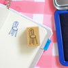 A wooden rubber stamp with a chair design, shown next to an open notebook with a stamped blue chair drawing and a blue ink pad on a pink and white checkered background.