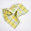 A folded kitchen towel with a retro pattern of yellow flowers and green stripes on a white background, photographed on a clean white surface.