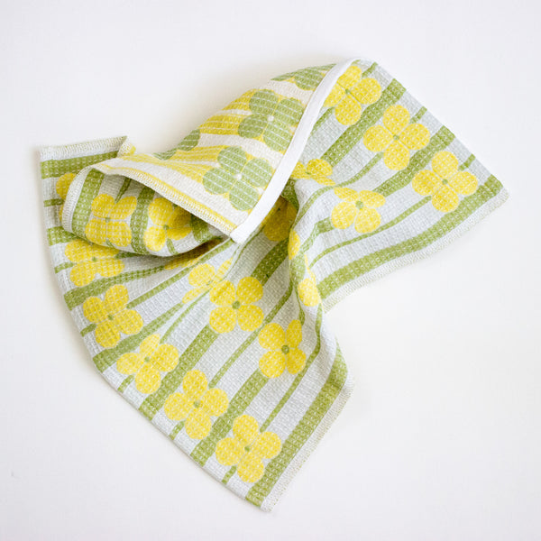A folded kitchen towel with a retro pattern of yellow flowers and green stripes on a white background, photographed on a clean white surface.
