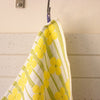 A close-up of a waffle-weave kitchen towel with a yellow flower pattern on a white background, hanging from a silver hook against a cream-coloured wall.