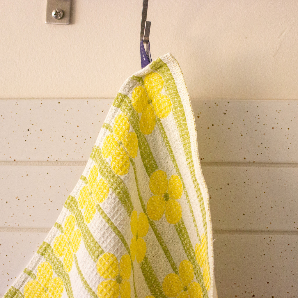 A close-up of a waffle-weave kitchen towel with a yellow flower pattern on a white background, hanging from a silver hook against a cream-coloured wall.