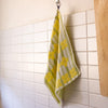 A yellow, green and white patterned kitchen towel hangs from a metal hook on a white tiled wall.