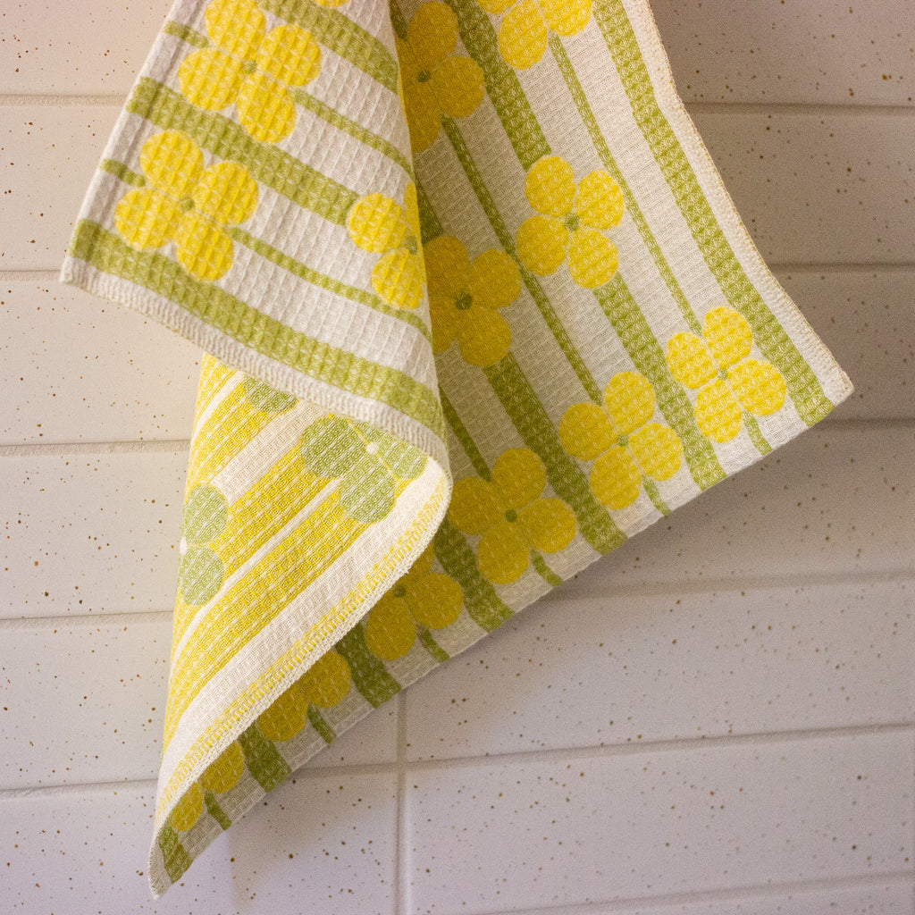 A folded kitchen towel with a retro pattern of yellow flowers and green stripes on a white background, hanging against a white paneled wall.