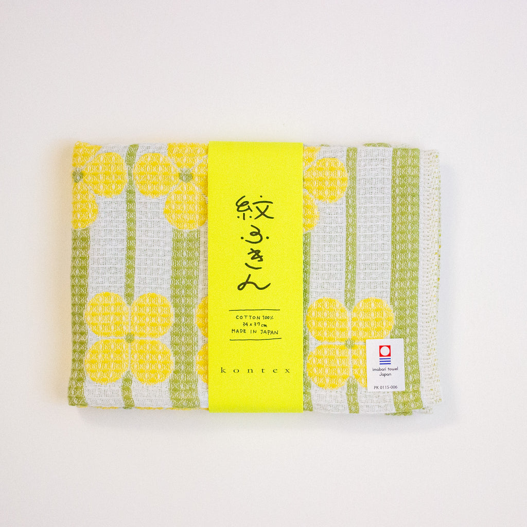 A folded Japanese cotton tea towel with a yellow and green floral pattern, wrapped in a bright yellow paper band with Japanese calligraphy and a Kontex brand label, set against a plain white background.