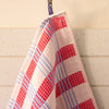 A close-up of a waffle-weave kitchen towel with a red, white, and light blue plaid pattern, hanging from a metal hook against a cream-coloured paneled wall.
