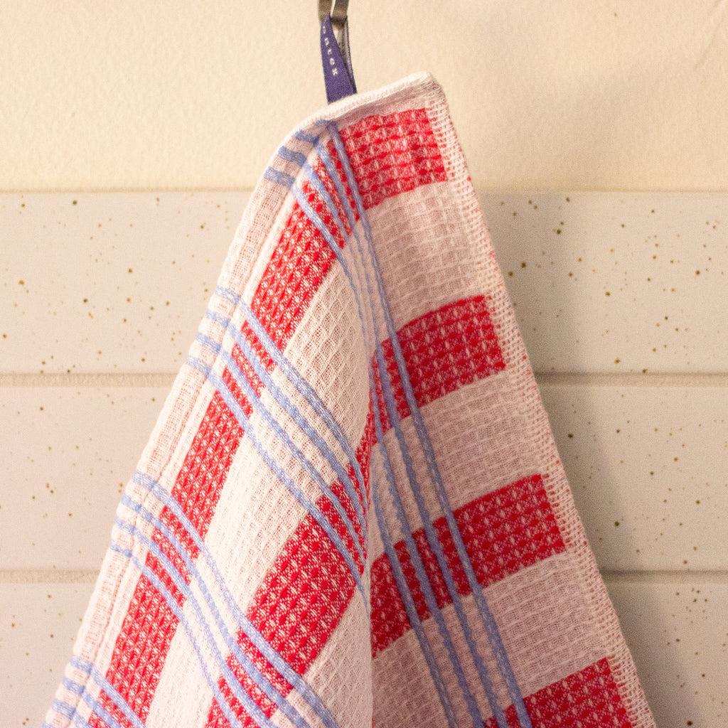 A close-up of a waffle-weave kitchen towel with a red, white, and light blue plaid pattern, hanging from a metal hook against a cream-coloured paneled wall.