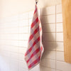 A red, white, and light blue plaid kitchen towel hanging from a metal hook on a white tiled wall.