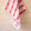 A white waffle-weave kitchen towel with a red and light blue plaid pattern, hanging against a light beige wood-paneled wall.