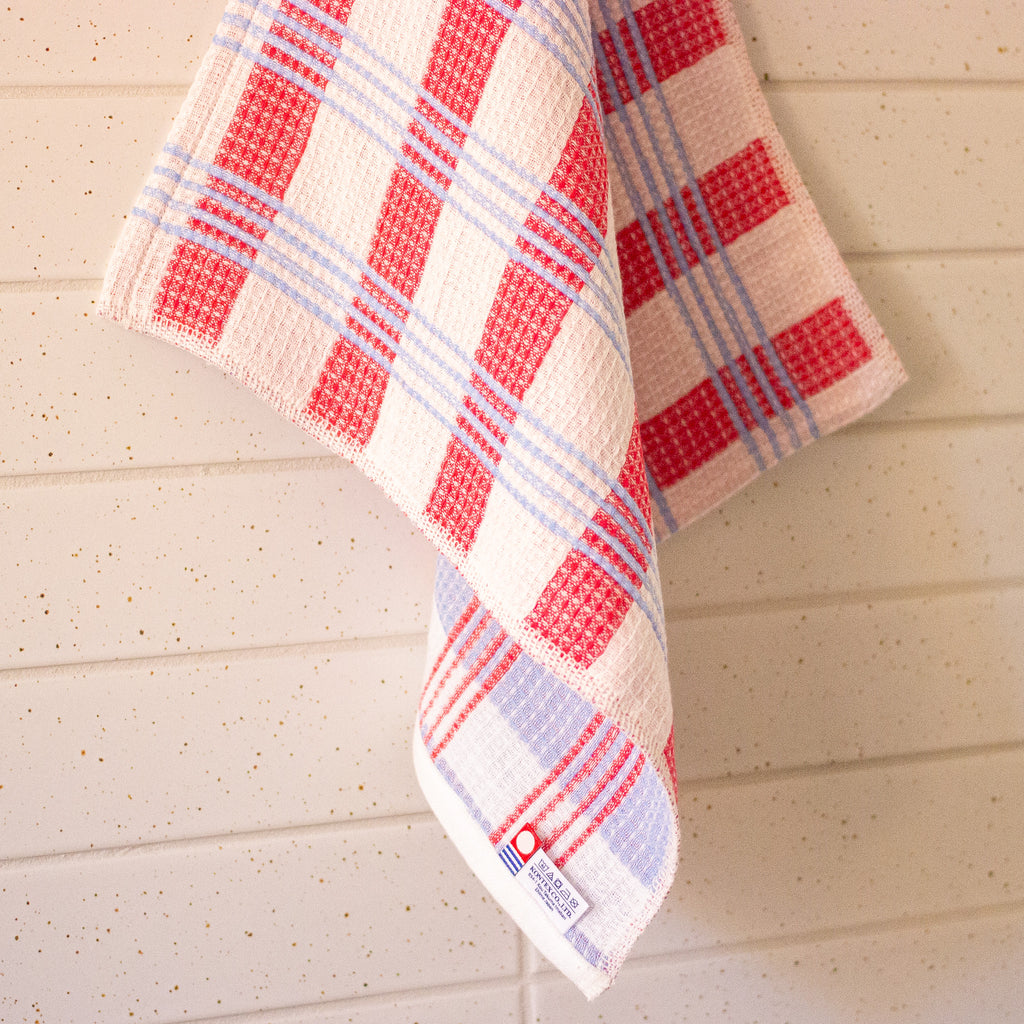 A white waffle-weave kitchen towel with a red and light blue plaid pattern, hanging against a light beige wood-paneled wall.