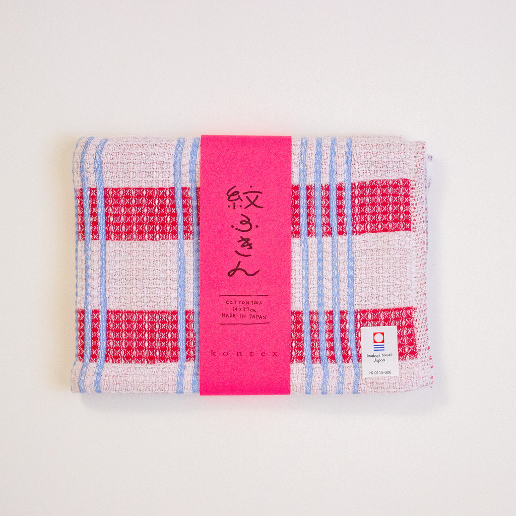 A folded cotton tea towel with a red, white, and light blue plaid pattern, wrapped in a bright pink paper band with Japanese text and a small white label.