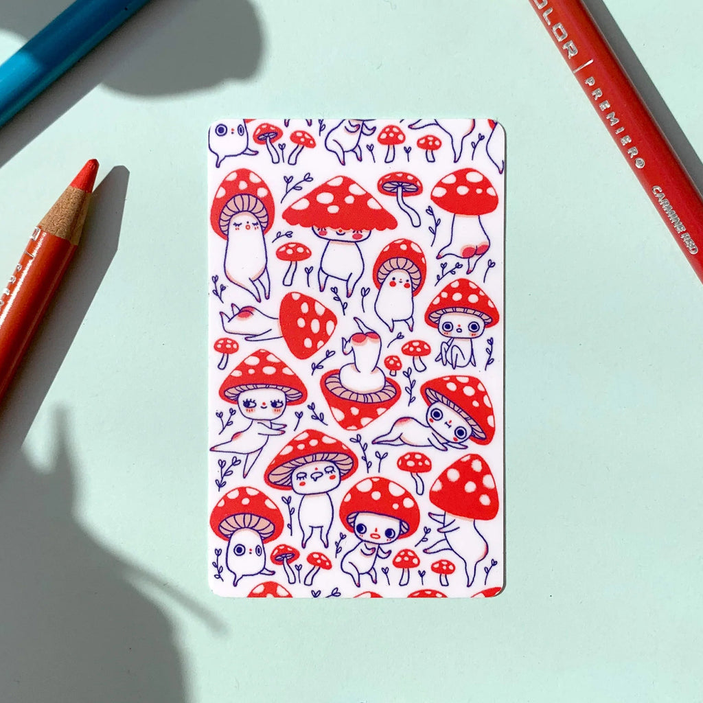 A rectangular sticker featuring a repeating pattern of cute red and white mushrooms with small cartoon figures, placed on a pale mint background with coloured pencils around it.