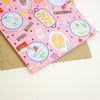 A pink greeting card with a whimsical food-themed illustration pattern, shown partially open to reveal a brown kraft envelope behind it on a light cream background.