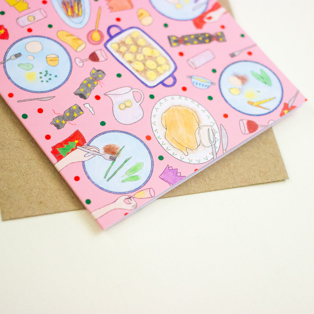A pink greeting card with a whimsical food-themed illustration pattern, shown partially open to reveal a brown kraft envelope behind it on a light cream background.