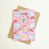 A pink greeting card with a playful food-themed illustration and the word 'CHEERS!' at the top, shown with a brown kraft envelope on a light cream background.