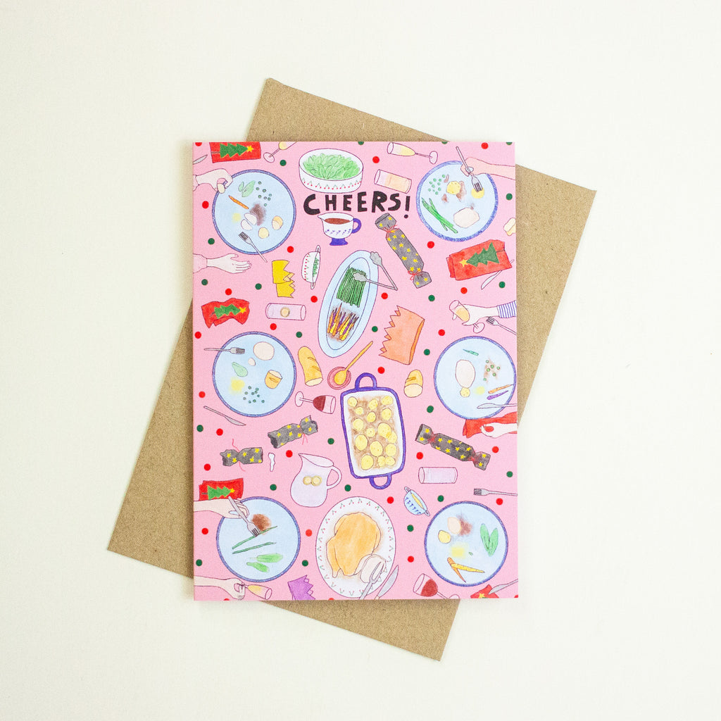 A pink greeting card with a playful food-themed illustration and the word 'CHEERS!' at the top, shown with a brown kraft envelope on a light cream background.