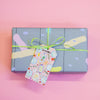 A rectangular gift box wrapped in pastel abstract-patterned paper, tied with a lime green ribbon and a decorative 'Cheers!' gift tag, set against a solid pink background.