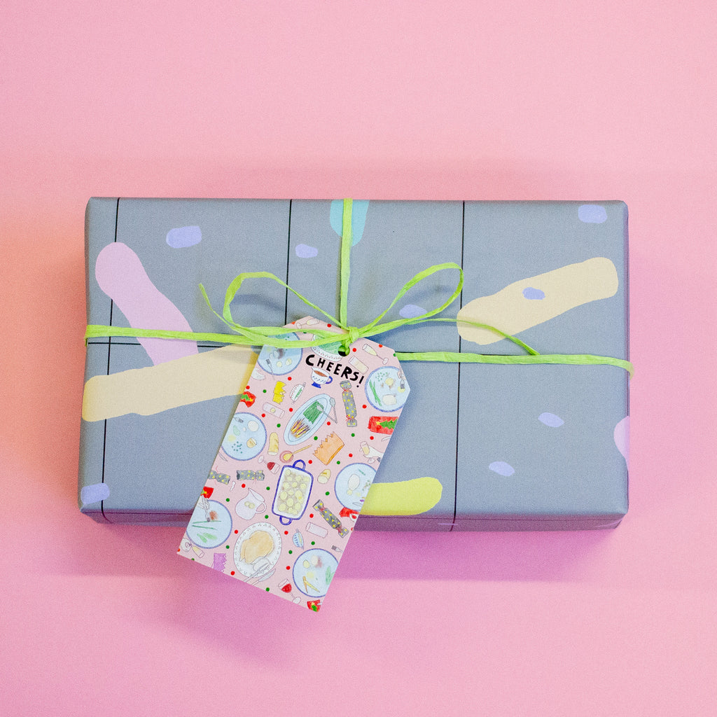 A rectangular gift box wrapped in pastel abstract-patterned paper, tied with a lime green ribbon and a decorative 'Cheers!' gift tag, set against a solid pink background.