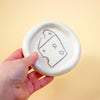 Hand holding a small white ceramic dish with a cheese illustration on a yellow background