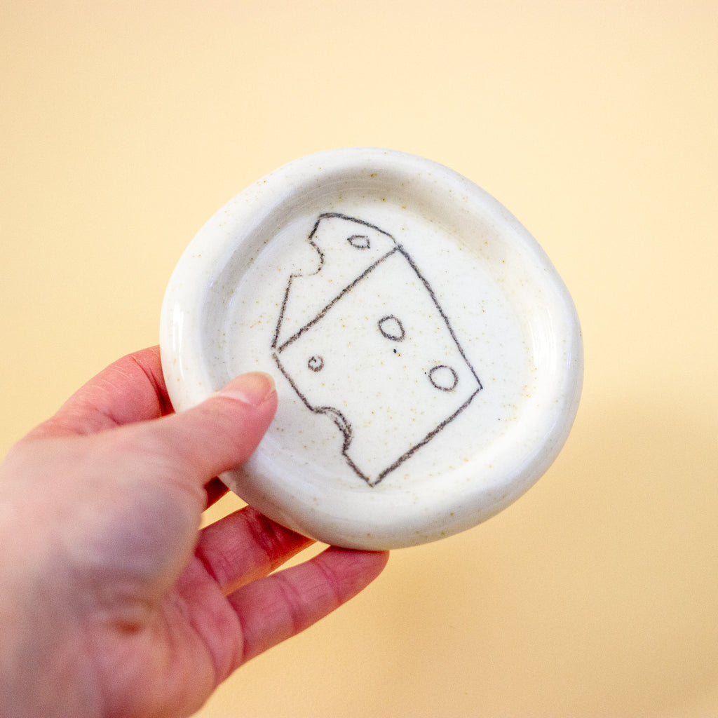 Hand holding a small white ceramic dish with a cheese illustration on a yellow background