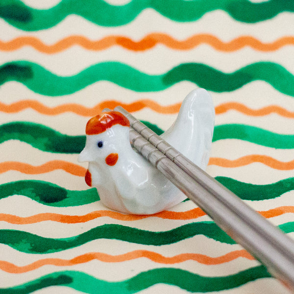 White ceramic chicken with red comb holding chopsticks against a wavy green and orange patterned background