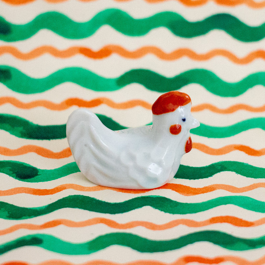 White ceramic chicken figurine on a wavy striped background with green and orange lines.