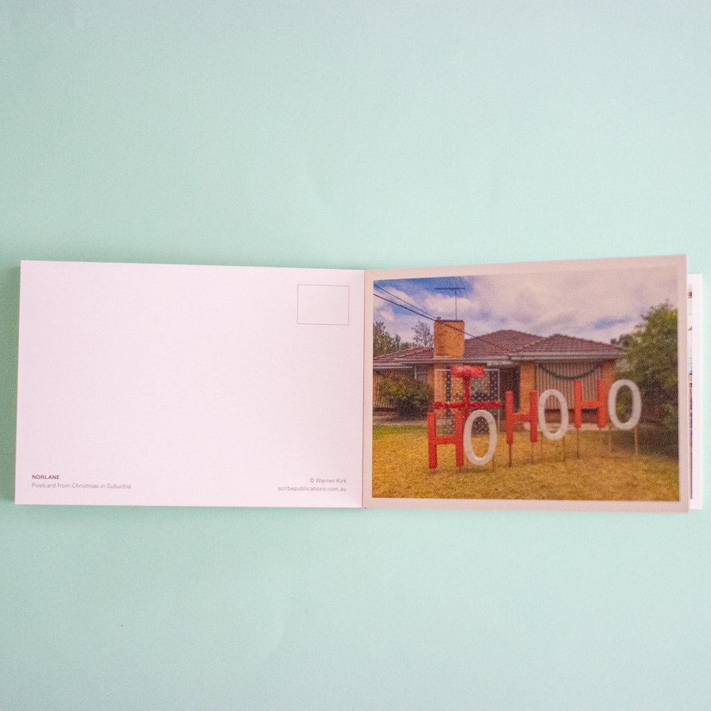 An open postcard album laid flat on a light mint green background. The left page is blank with a postcard back design, and the right page shows a printed photograph of a house with large red and white 'HO HO HO' letter signs in front.