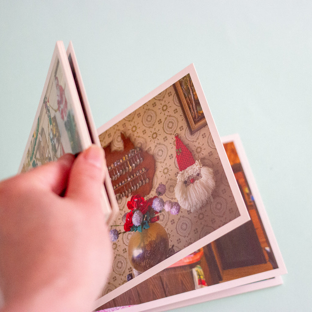 A hand holds a stack of printed postcards against a pale blue background. The top card shows a cosy indoor scene with a small gnome, a vase of flowers, and patterned wallpaper.