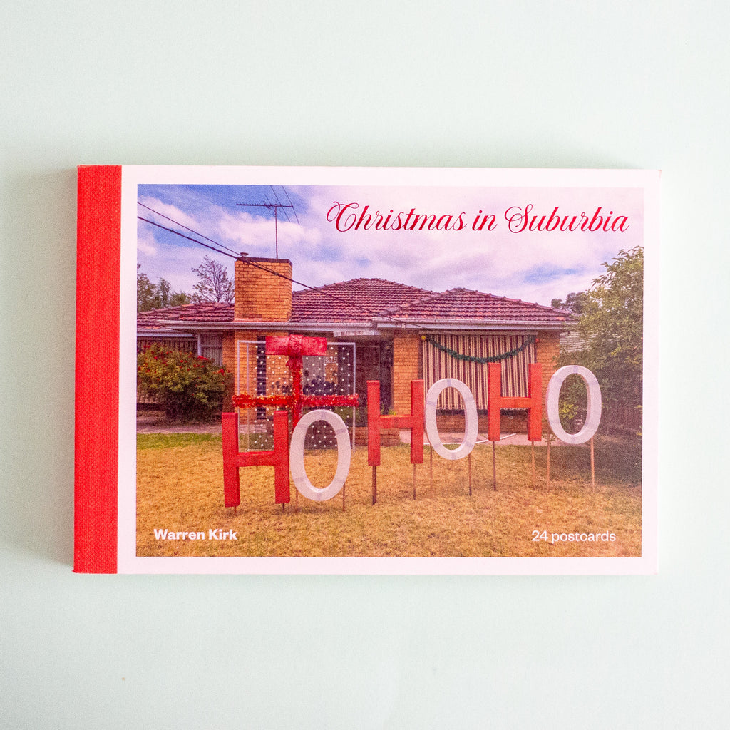 A set of postcards titled "Christmas in Suburbia" by Warren Kirk, shown standing upright against a pale mint background. The package has a red spine/border and the front features a photograph of a suburban house with large red and white "HOHOHO" lettering in the front yard.