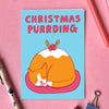 A square greeting card with a light blue background, featuring a cartoon orange cat curled up inside a Christmas pudding with the text 'CHRISTMAS PURRDING' in pink. The card is styled in a flat-lay on a pink background with pens, paper clips, and a patterned pen.