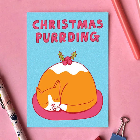 A square greeting card with a light blue background, featuring a cartoon orange cat curled up inside a Christmas pudding with the text 'CHRISTMAS PURRDING' in pink. The card is styled in a flat-lay on a pink background with pens, paper clips, and a patterned pen.