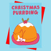 A square greeting card or print with the phrase “CHRISTMAS PURRDING” in pink letters above a sleeping orange-and-white cat curled up like a pudding on a pink plate, set against a light blue background with red envelope behind it.