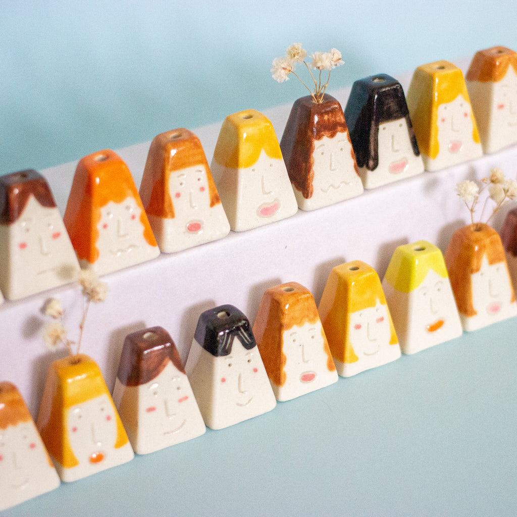 A row of small ceramic face-shaped vases with different hair colours, arranged on a white shelf against a light blue background.