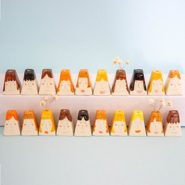 A set of small decorative ceramic volcano-shaped objects arranged in two rows on a pale blue background. Each piece has a cream base with painted faces and hair in orange, yellow, brown, and black, with a few small dried flowers placed in front of some pieces.