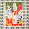 An art print featuring a whimsical illustration of white rabbits sitting at small tables with red stools, drinking from glasses and reading a newspaper. 