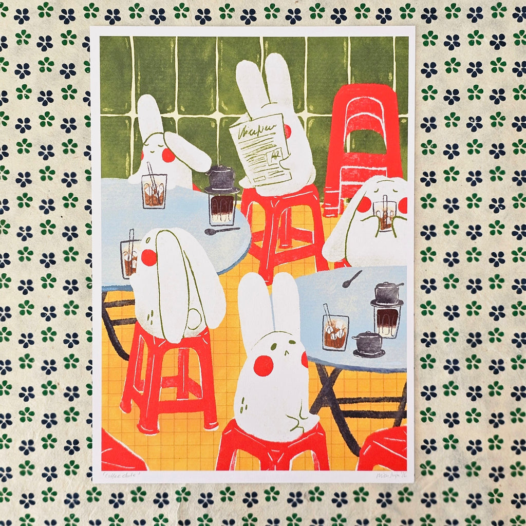 An art print featuring a whimsical illustration of white rabbits sitting at small tables with red stools, drinking from glasses and reading a newspaper. 