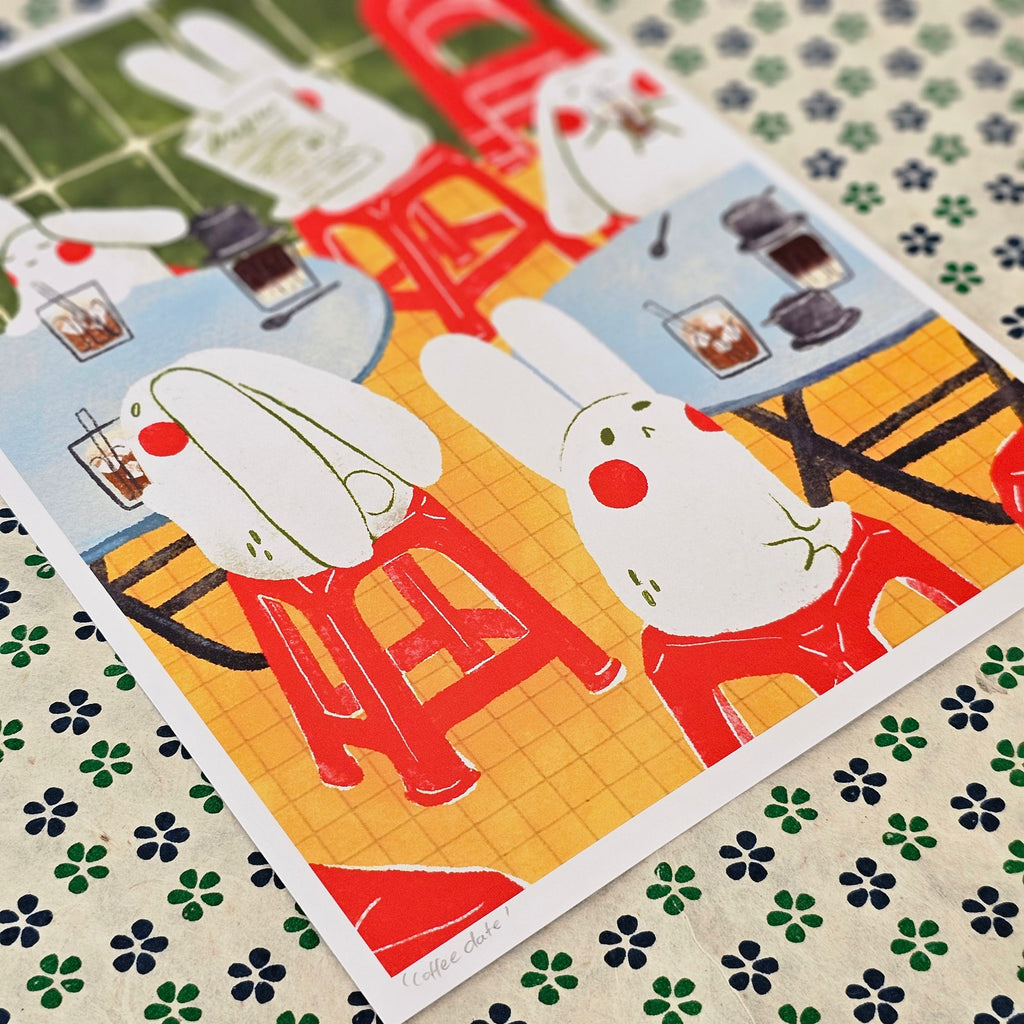 An art print featuring a whimsical illustration of white rabbits sitting at red tables with drinks, placed diagonally on a patterned fabric background.