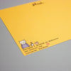 A close-up of a yellow postcard placed on a grey surface. The card features a small illustrated cat in a striped shirt, a coffee cup, an envelope, and printed text including 'Designed & printed in Australia', 'www.shuhlee.com'. 