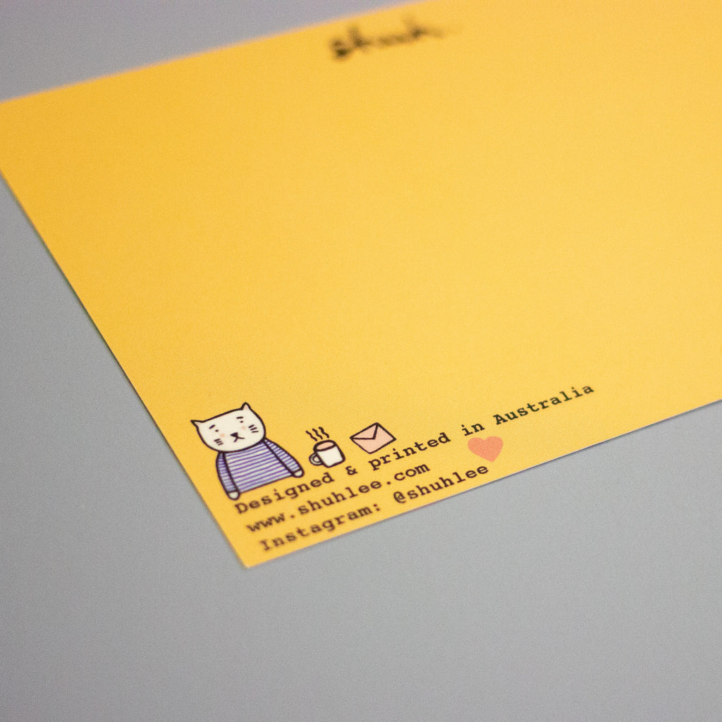 A close-up of a yellow postcard placed on a grey surface. The card features a small illustrated cat in a striped shirt, a coffee cup, an envelope, and printed text including 'Designed & printed in Australia', 'www.shuhlee.com'. 