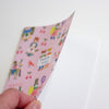 A hand holding a pink notebook with a colourful cartoon pattern, against a plain white background.