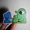A hand holding a die-cut sticker featuring a green cartoon figure sitting at a blue computer, against a plain light background.
