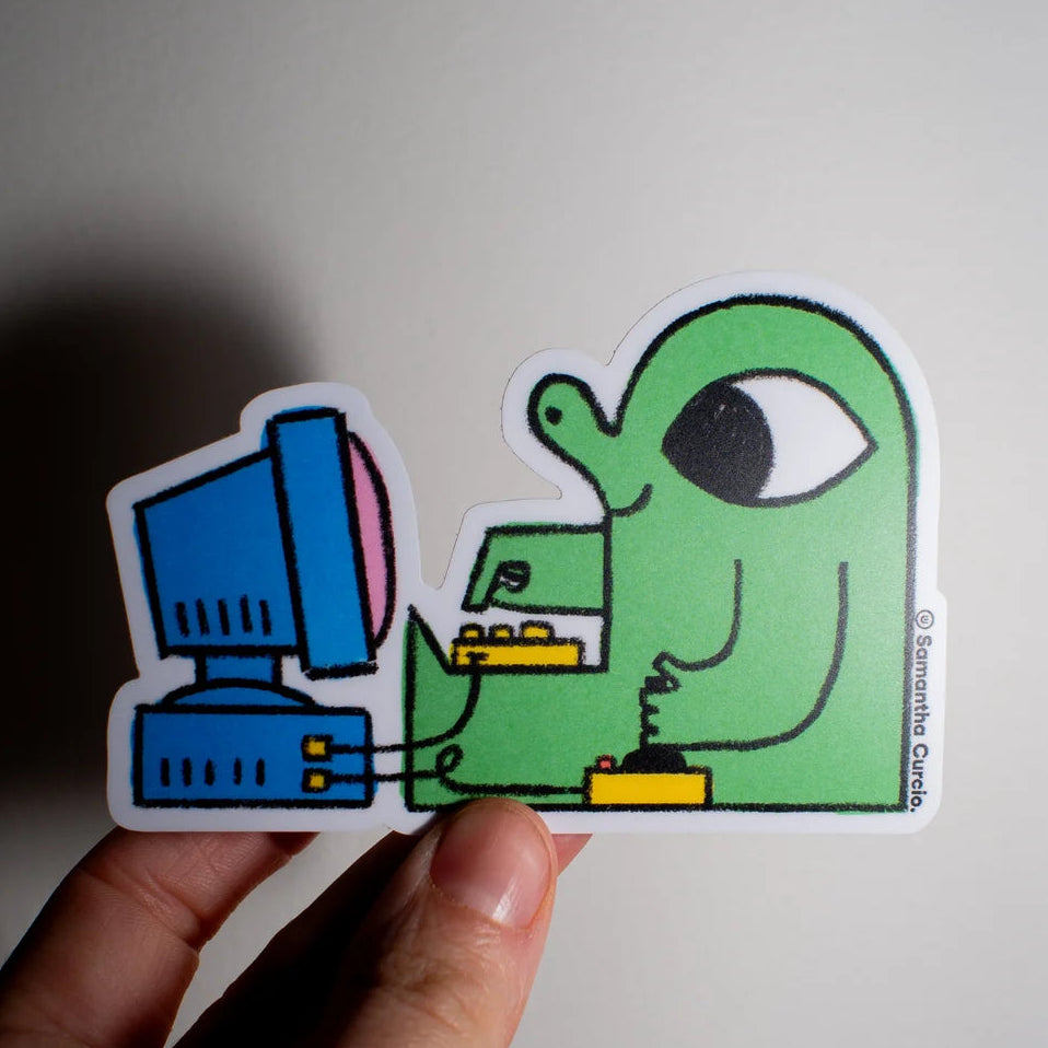A hand holding a die-cut sticker featuring a green cartoon figure sitting at a blue computer, against a plain light background.