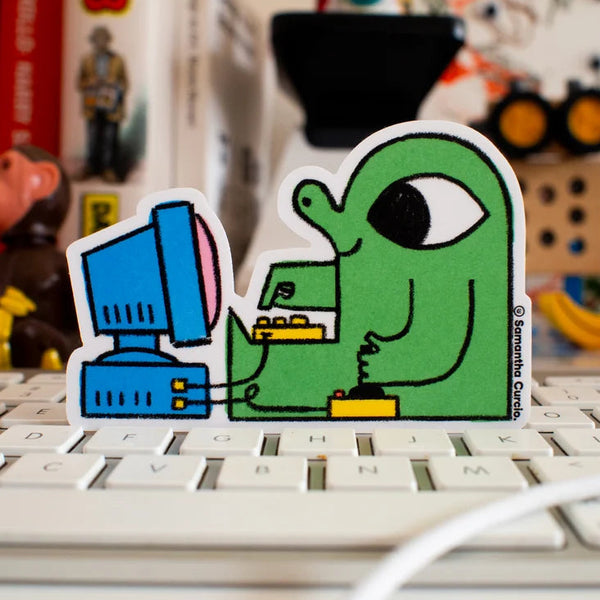 A die-cut sticker featuring a green cartoon figure typing on a blue computer, placed on a white keyboard with a blurred desk background.