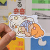 A hand holding a sticker of a girl inside a shell against a colourful background. 