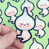 Hand holding a cute cartoon-style garlic character sticker with a pattern of similar stickers in the background.