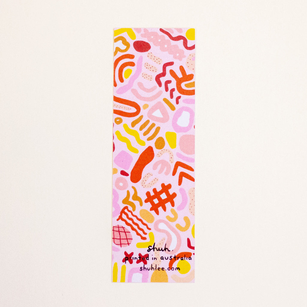 A tall, narrow rectangular bookmark featuring a playful abstract pattern in pink, orange, yellow, and red on a white background, with the text 'shuh.' and 'shuhlee.com' printed at the bottom.