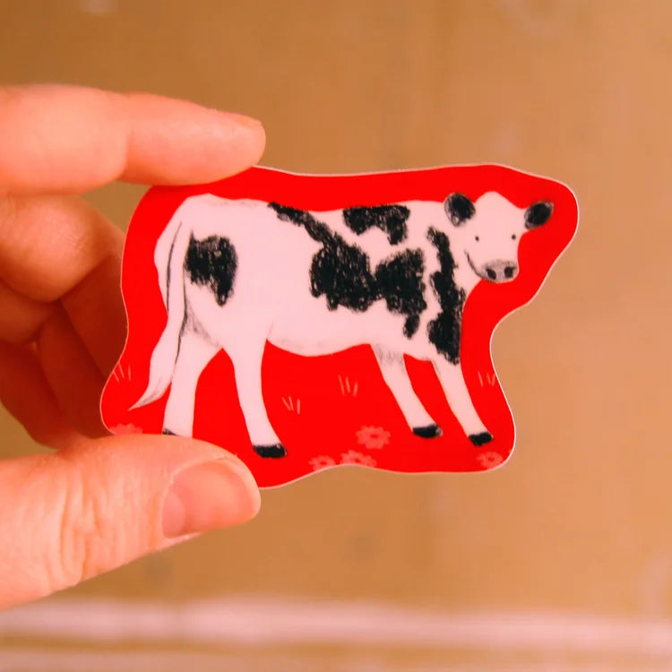 A hand holding a die-cut sticker featuring a black and white cow on a bright red background, set against a soft-focus warm brown backdrop.