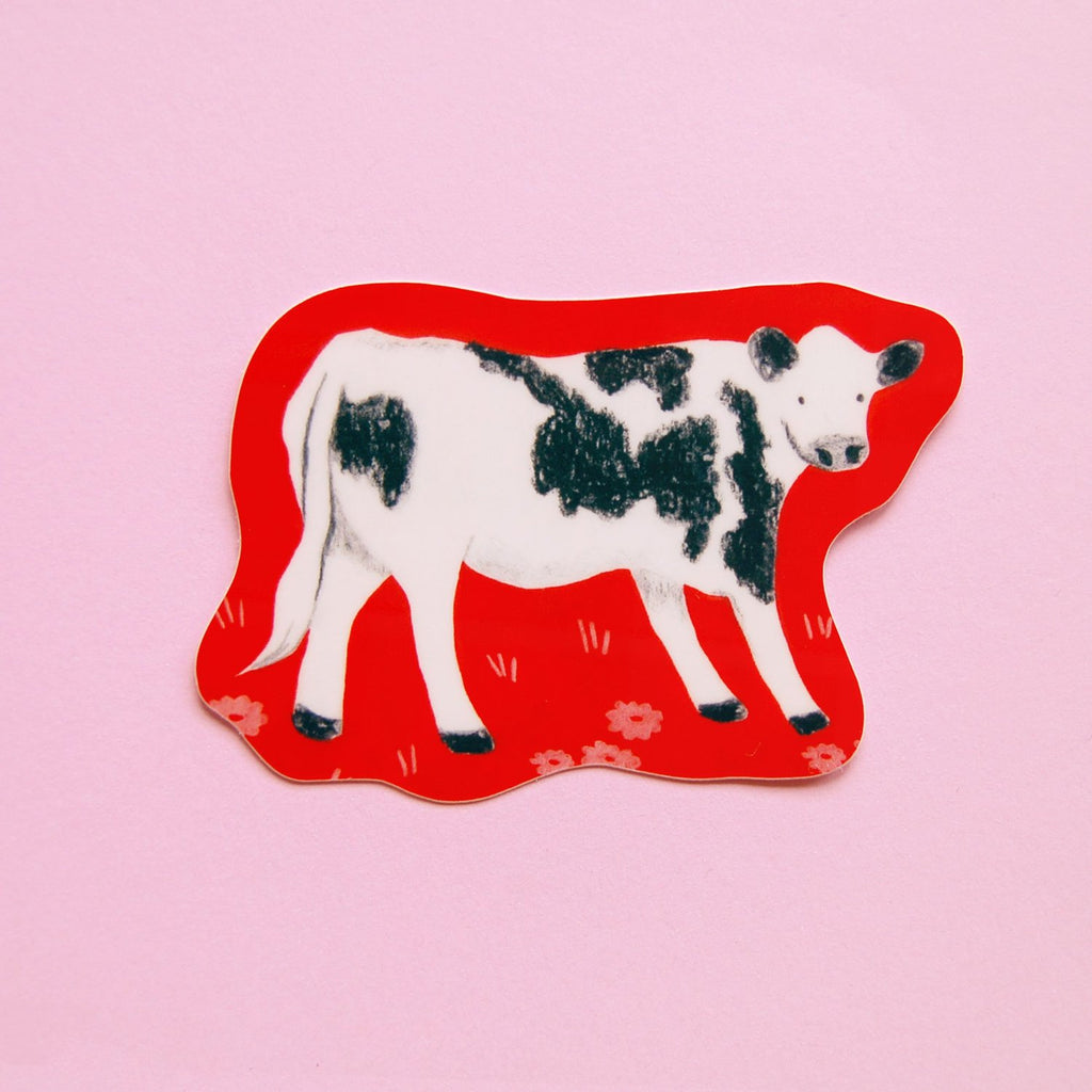 A die-cut sticker or magnet featuring a black and white cow standing on a bright red background with small white flowers, shown on a solid pink surface.