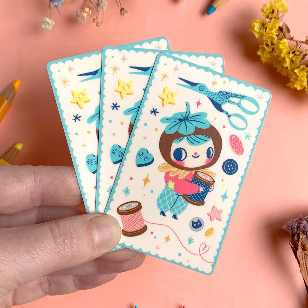 Hand holding a card with a cartoon character on a pink background with pencils and flowers.