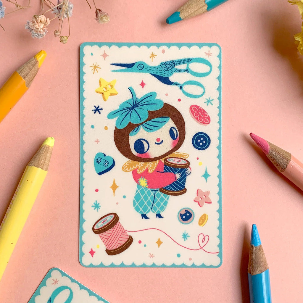 A colourful illustrated card or sticker sheet featuring a cartoon girl with a blue bow, surrounded by sewing-themed doodles like scissors, buttons, and thread spools, arranged on a pink surface with coloured pencils and dried flowers.