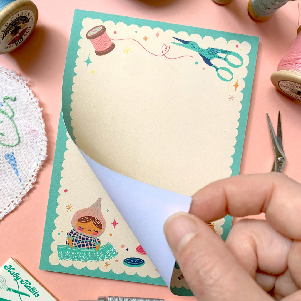 A hand peels back a page from a decorative stationery notepad. The notepad has a scalloped mint border, cream writing area, and whimsical sewing-themed illustrations including scissors, a spool, and a fairy.